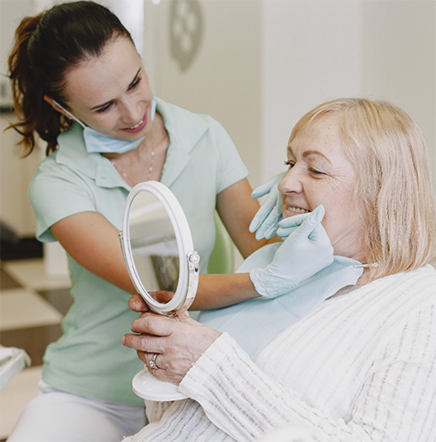 Senior woman in chair checking smile in mirror while dentist watches
